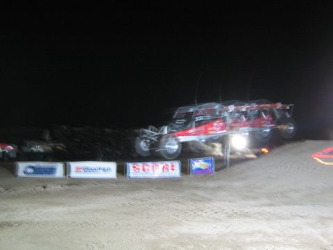 1990 Ford Bronco - red and black off-road race buggy flies over a floodlit dirt jump at night above SCORE and BFGoodrich banners.