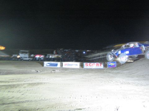 1990 Ford Bronco - blue off-road race vehicle motion-blurred over a dirt course at night, with support trucks and SCORE banners in the background.