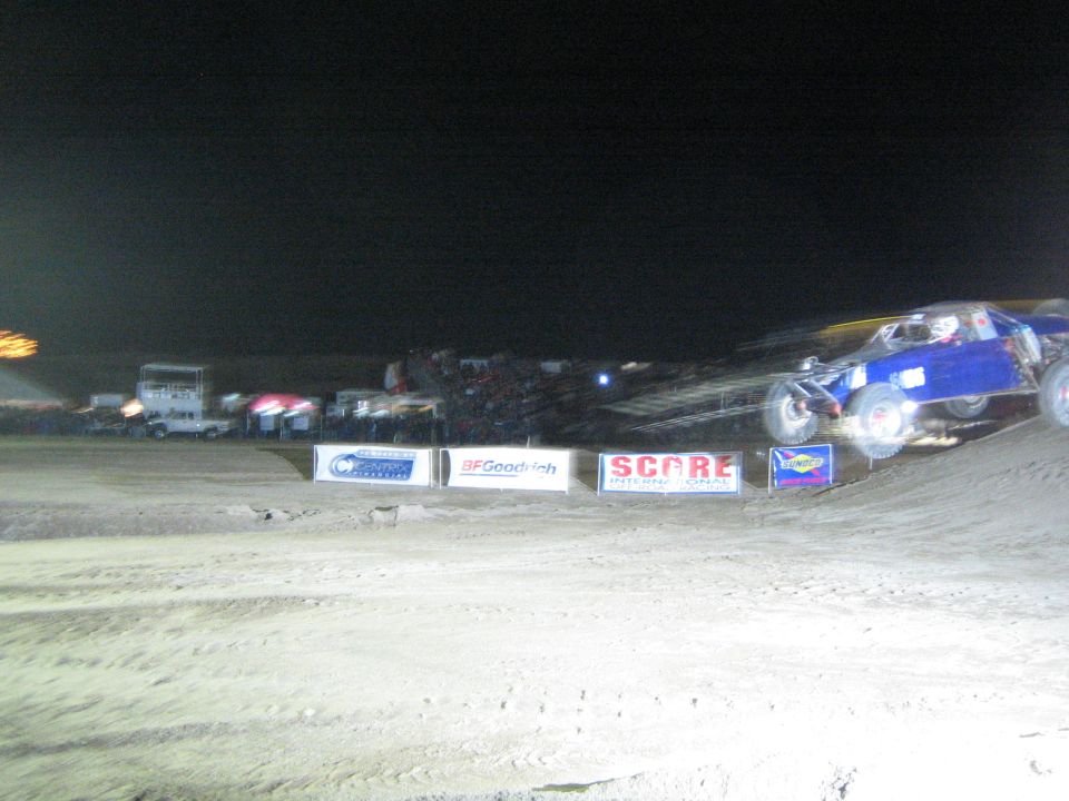 Blue off-road race vehicle on a dirt course at night with banners