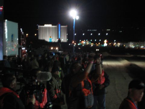 1990 Ford Bronco - crowd of spectators in orange-trimmed jackets gathers at night under a floodlight, a large illuminated hotel rising in the distance.