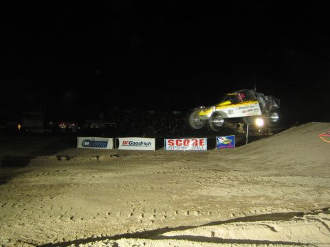 1990 Ford Bronco - yellow and black off-road race truck crests a large floodlit dirt jump at night above SCORE and BFGoodrich banners.