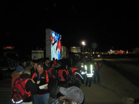 1990 Ford Bronco - Spectators in red jackets and a safety official in hi-vis gather at night beside a large outdoor LED video screen, city lights glowing behi…