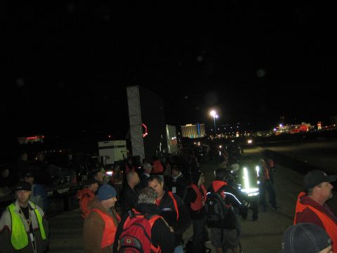 1990 Ford Bronco - crowd of spectators, some in orange safety vests, stands at night near a vehicle with floodlights and distant city lights in the background.