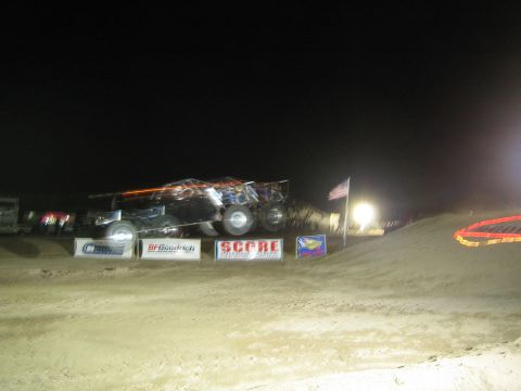 1990 Ford Bronco - black and orange off-road race vehicle crests a dirt jump at night beside an American flag and SCORE banners under floodlights.