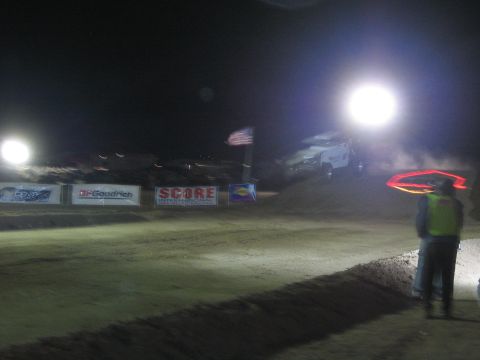 1990 Ford Bronco - course official in a hi-vis vest watches as a race vehicle crests a floodlit dirt jump at night beside an American flag and SCORE banners.