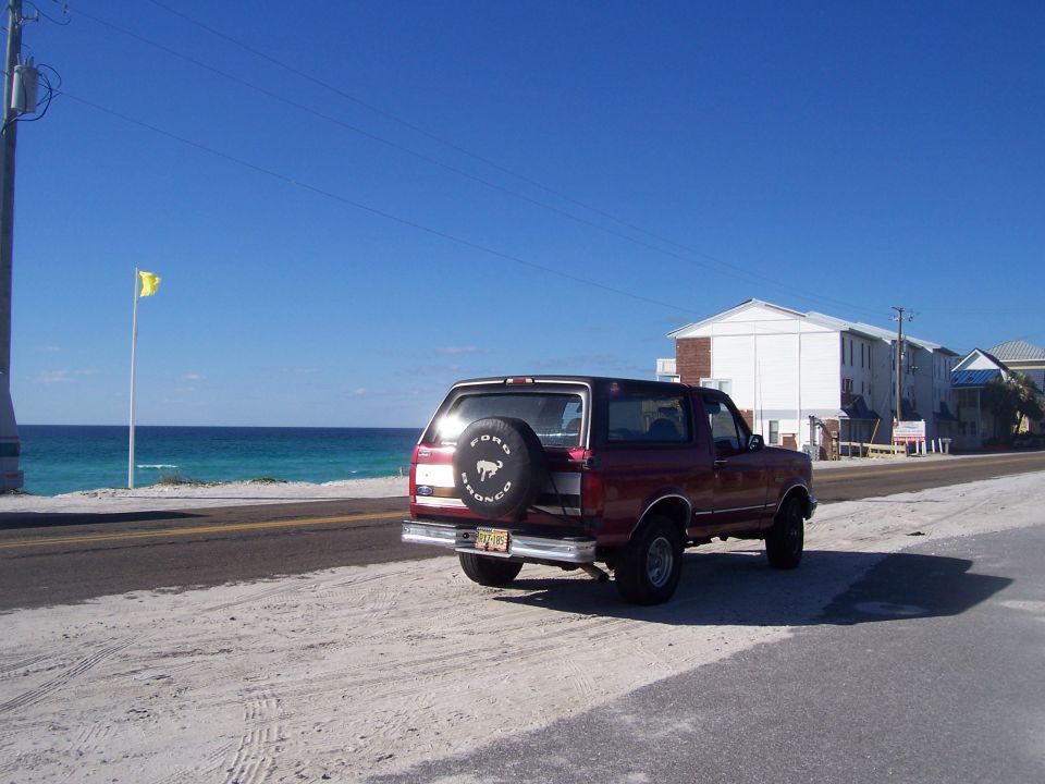 Red Ford Bronco parked on a sandy road next to the ocean. Yellow flag on a pole