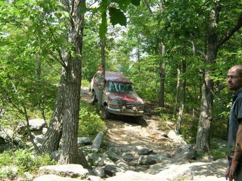 1995 Ford Bronco - A red SUV navigates a challenging off-road path through a dense forest.