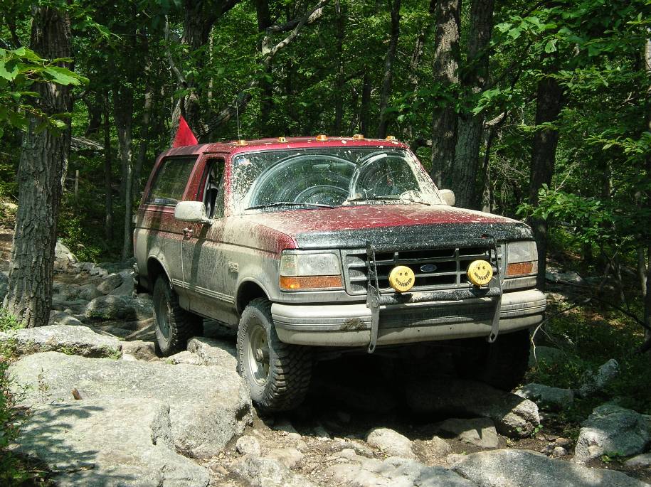 Muddy red and tan Ford Bronco on rocks with yellow smiley-face driving lights