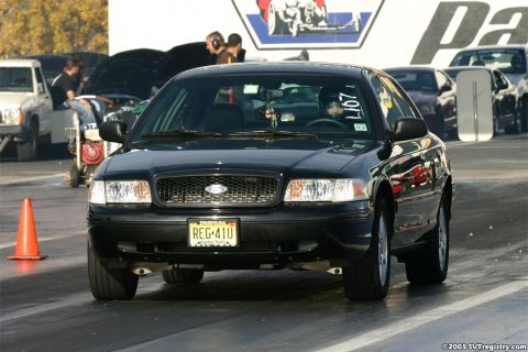 2004 Ford Crown Victoria - A black 2004 Ford Crown Victoria is lined up on a drag strip, with other vehicles and people in the background.