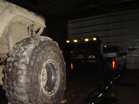 1992 Ford Bronco - A large, mud-covered tire dominates the foreground, obscuring the view of a tow truck and another vehicle in the dimly lit background.