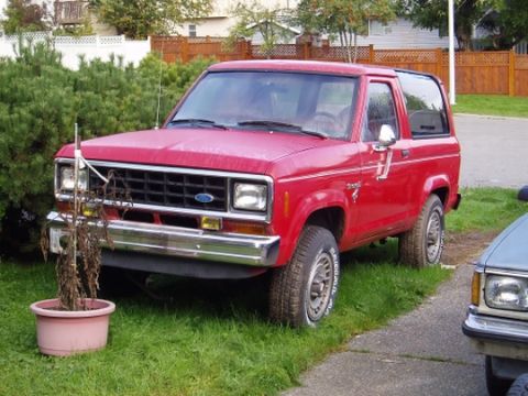 1985 Ford Bronco II - A red Ford Bronco II is parked in a residential driveway, partially obscured by bushes and another vehicle.
