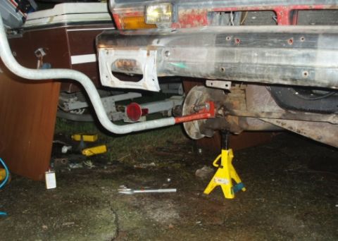 1985 Ford Bronco II - Close-up view of a vehicle's rear axle and suspension components being worked on, supported by a yellow jack stand.