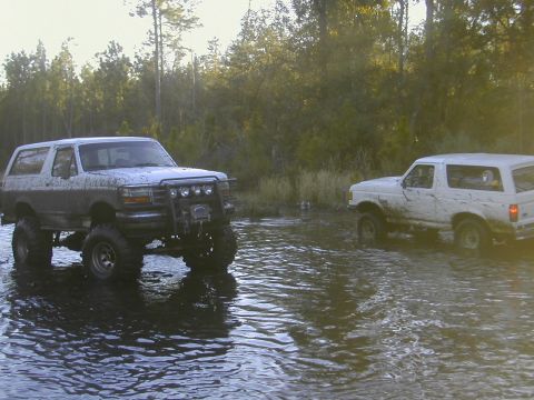 1993 Ford Bronco - Two muddy Ford Broncos navigate through a watery terrain surrounded by trees.