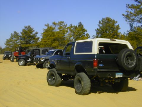1993 Ford Bronco - A line of off-road vehicles, including a dark blue Ford Bronco, are parked on a sandy surface under a clear blue sky.