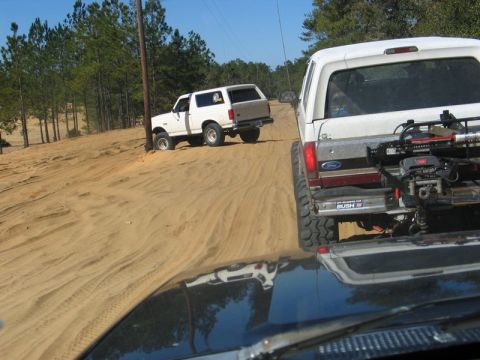 1995 Ford Bronco - Two Ford Broncos navigate a sandy off-road path surrounded by trees and a utility pole.