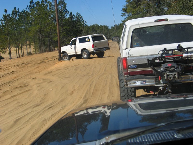 Two white Ford Broncos driving on a sandy trail. One is in front, seen from the rear, with a rack on the back. The other is further up the trail