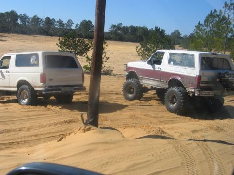 1995 Ford Bronco - Two Ford Broncos tackle sandy terrain, one lifted high with aggressive off-road tires.