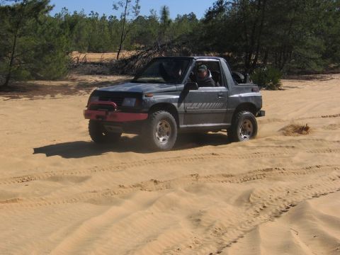 1995 Ford Bronco - A dark gray convertible SUV navigates sandy terrain, kicking up dust as it drives.