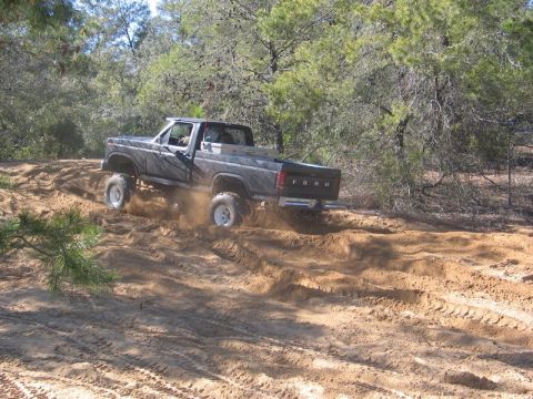 1995 Ford Bronco - A lifted Ford pickup truck navigates a sandy off-road path amidst trees.