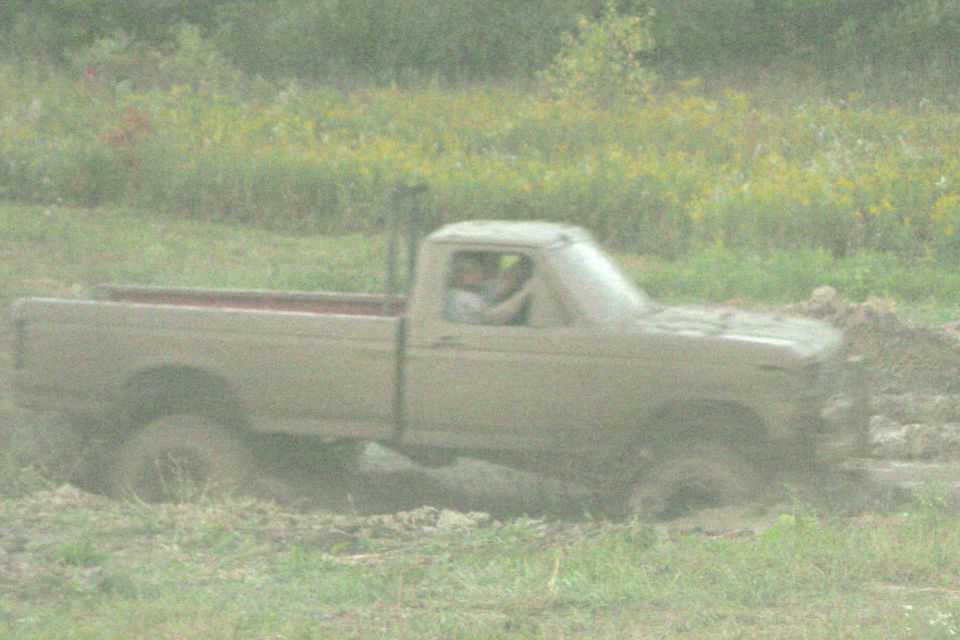 Mud-covered tan Ford pickup stuck in deep mud bog, side view, two people in cab