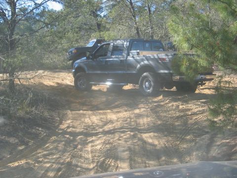 1995 Ford Bronco - A dark pickup truck navigates a sandy trail, with another vehicle visible in the background.
