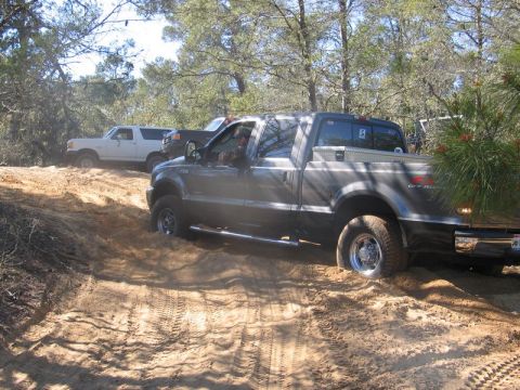 1995 Ford Bronco - A gray pickup truck navigates sandy terrain off-road, with other vehicles visible in the background.