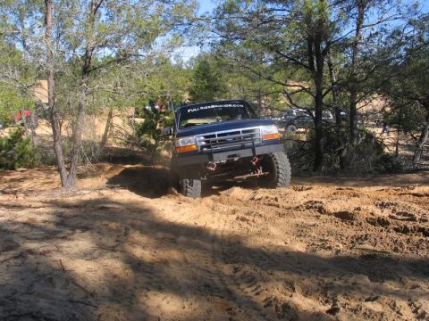 1995 Ford Bronco - A dark blue Ford Bronco navigates a sandy terrain with trees in the background.