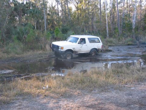 1995 Ford Bronco - A white Ford Bronco navigates a muddy trail, its tires splashing through the water.