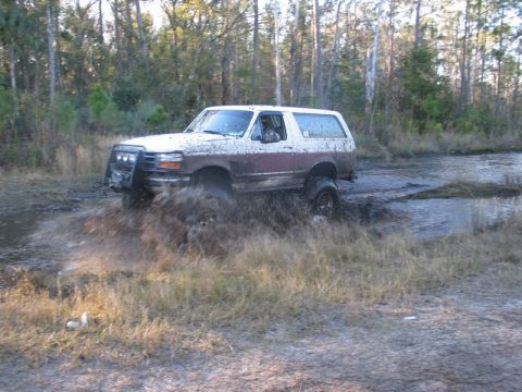1995 Ford Bronco - A white and brown 1995 Ford Bronco navigates a muddy trail, kicking up water and dirt.