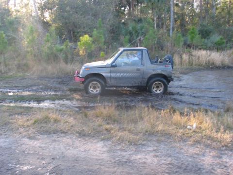 1995 Ford Bronco - Suzuki Samurai navigating a muddy trail with trees in the background.