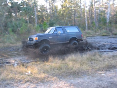 1995 Ford Bronco - A dark SUV kicks up a large spray of mud as it drives through a muddy area.