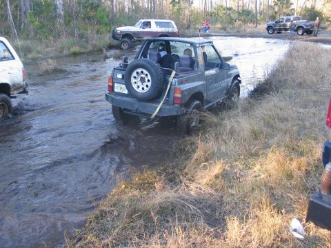 1995 Ford Bronco - Several off-road vehicles navigate a muddy terrain with trees in the background.