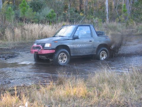 1995 Ford Bronco - A gray Suzuki Sidekick navigates a muddy trail, splashing water as it drives.