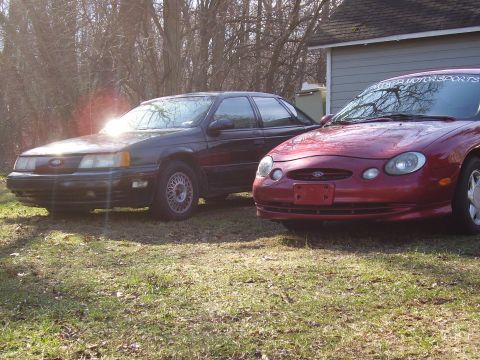 1997 Ford Taurus - Two Ford Taurus sedans are parked side-by-side in a grassy yard with trees in the background.