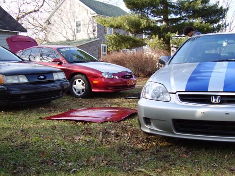1997 Ford Taurus - A red and silver 1997 Ford Taurus is parked next to other vehicles, with its hood open and a detached hood on the grass.