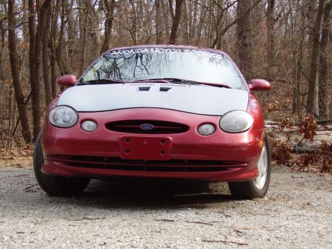 1997 Ford Taurus - This red car with a white hood is seen from the front on a gravel road surrounded by trees.