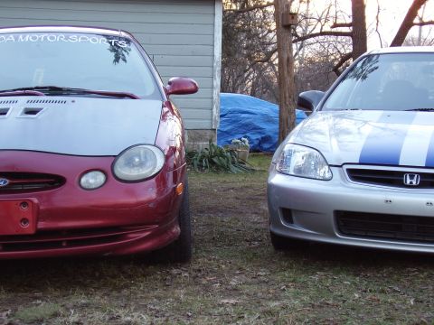 1997 Ford Taurus - Two cars are parked in front of a building, one red with a silver hood and the other silver with blue racing stripes.