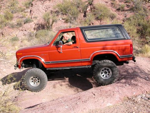 1984 Ford Bronco - A man drives an orange Ford Bronco through a dusty, rocky terrain with sparse vegetation.