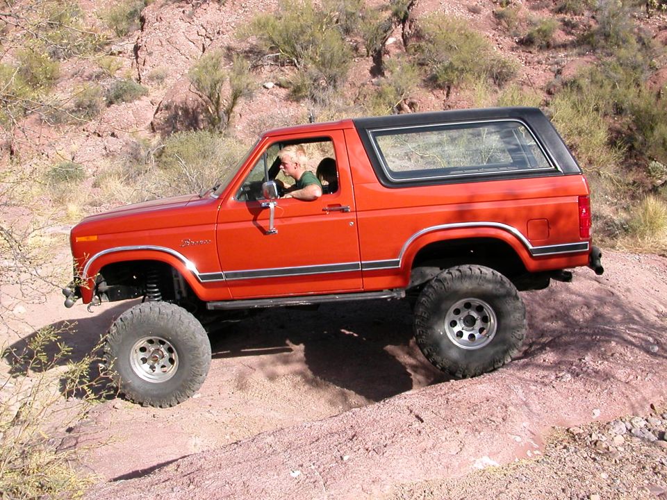 Orange Ford Bronco with lifted suspension and large tires driving on a dirt trail in a desert landscape