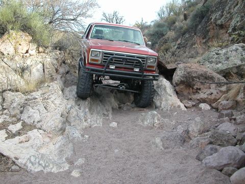 1984 Ford Bronco - This red Ford Bronco is tackling a challenging off-road course with large rocks and uneven terrain.