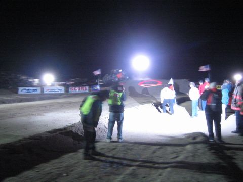 1990 Ford Bronco - Spectators and hi-vis officials line a floodlit dirt course at night as a race vehicle crests a jump near American flags and SCORE banners.