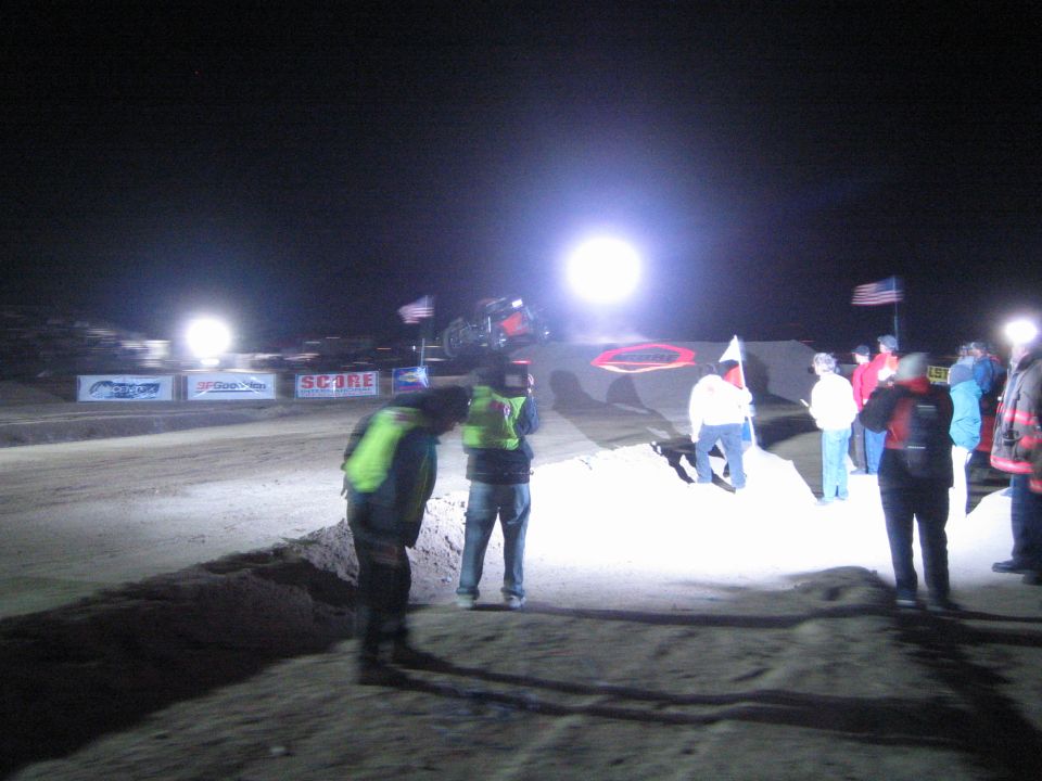 Spectators and officials watching a race vehicle on a floodlit dirt jump at night