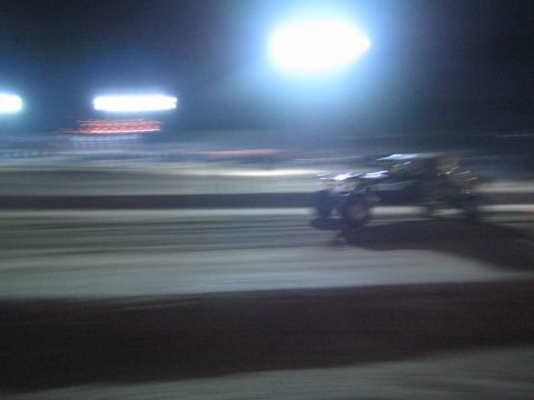 1990 Ford Bronco - race vehicle blurs across a dirt track at night, panned under bright floodlights with banners glowing in the background.
