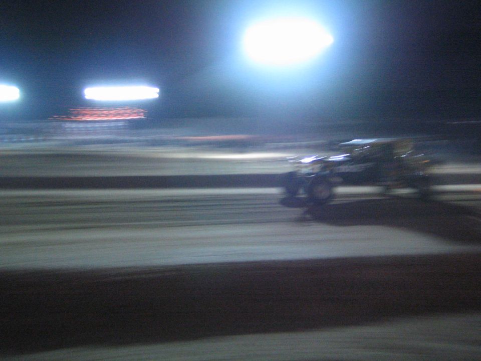 Motion-blurred race vehicle speeding across a dirt track at night under lights
