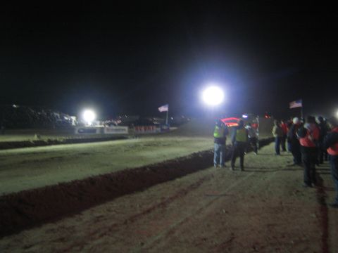 1990 Ford Bronco - Spectators and hi-vis officials line a floodlit dirt race course at night beside American flags and sponsor banners.