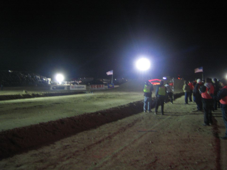 Spectators and officials lining a floodlit dirt race course at night