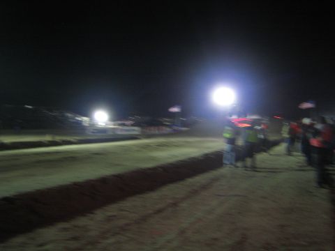 1990 Ford Bronco - blurry nighttime view of a dirt race course with bright floodlights, American flags, and spectators in hi-vis vests along the edge.