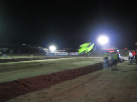 1990 Ford Bronco - green race vehicle streaks past hi-vis officials and spectators on a floodlit dirt track at night, headlights blazing.