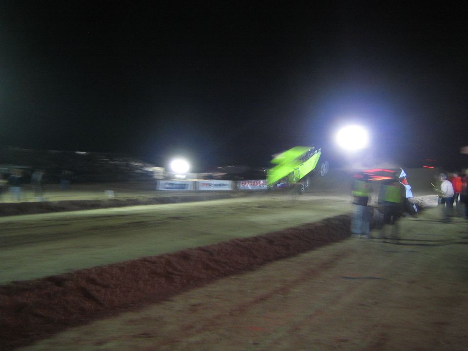 Green race vehicle speeding past spectators on a dirt track at night