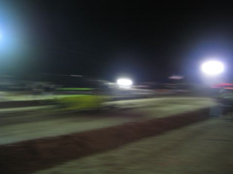 1990 Ford Bronco - heavily motion-blurred nighttime dirt race track lit by floodlights, with a faint green-lit race vehicle moving in the distance.